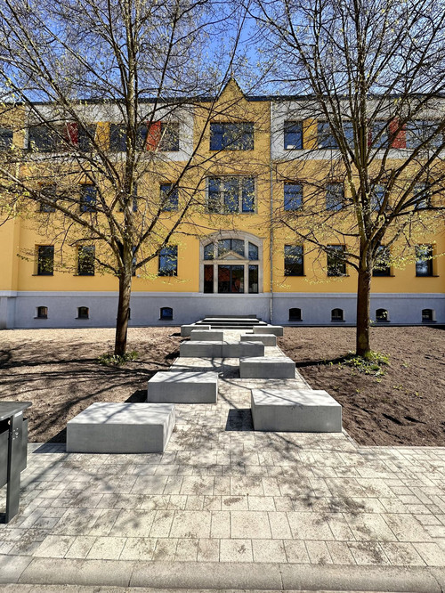 Blick auf den ehemaligen Haupteingang, der im Zuge der Komplettsanierung zu einem Deko-Fenster umfunktioniert wurde. Davor stehen Sitzgelegenheiten.